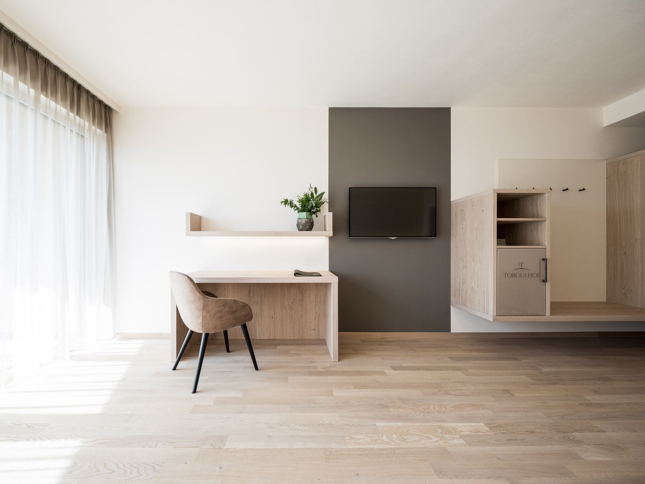 Heller, minimalistischer Arbeitsbereich mit Holzschreibtisch, beigem Stuhl, Fernseher an grauer Wand und bodentiefem Fenster mit Vorhängen.