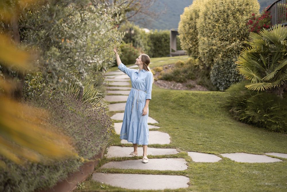 Frau im blauen Kleid berührt Blätter an einem Baum auf einem Gartenspfad.