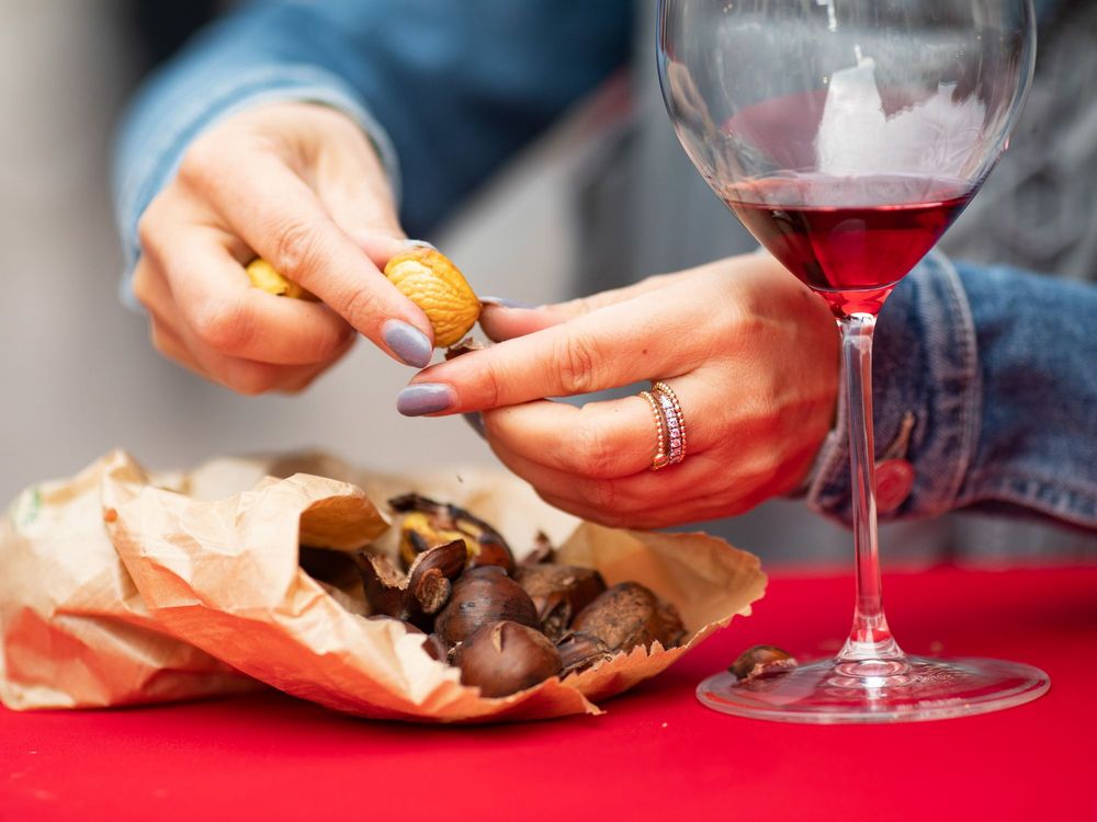 Person knackt Maroni, daneben ein Glas Rotwein auf rotem Tisch