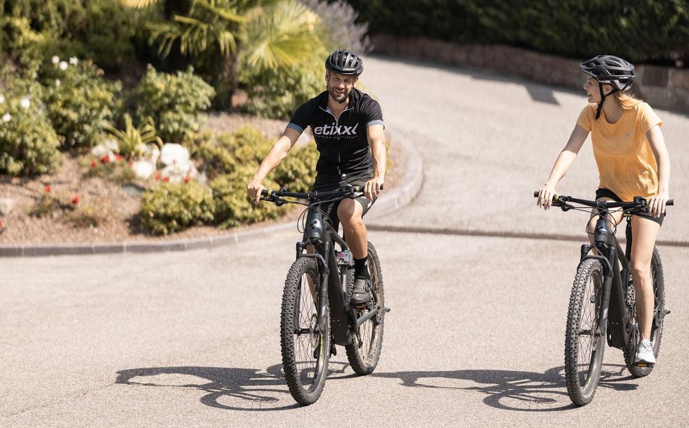 Zwei Personen fahren mit Helmen auf E-Bikes entlang einer Straße bei sonnigem Wetter.