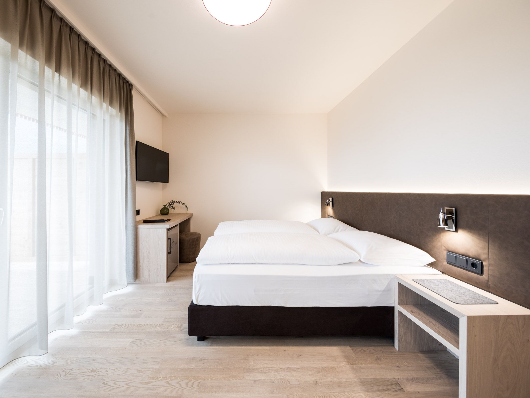 Modernes, schlicht eingerichtetes Schlafzimmer mit Doppelbett, hellem Holzboden und großen Fenstern mit Gardinen