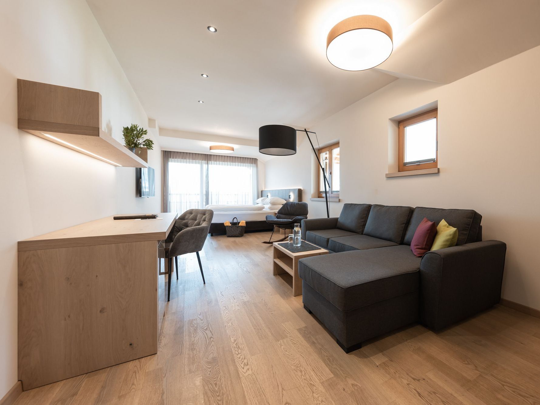 Modern eingerichtetes Wohnzimmer mit grauem Sofa, Schreibtisch und Bett im Hintergrund