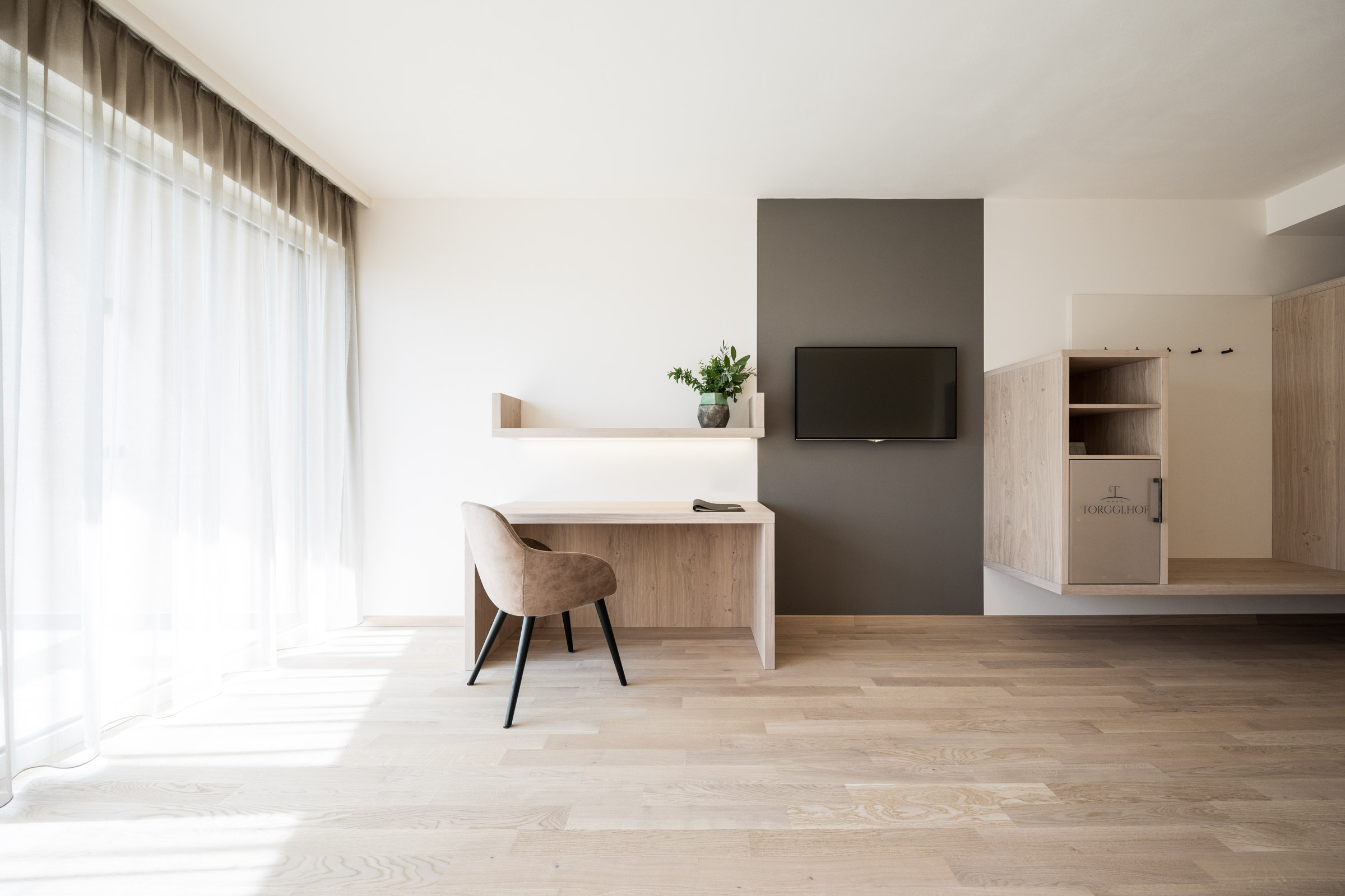 Heller, minimalistischer Arbeitsbereich mit Holzschreibtisch, beigem Stuhl, Fernseher an grauer Wand und bodentiefem Fenster mit Vorhängen.