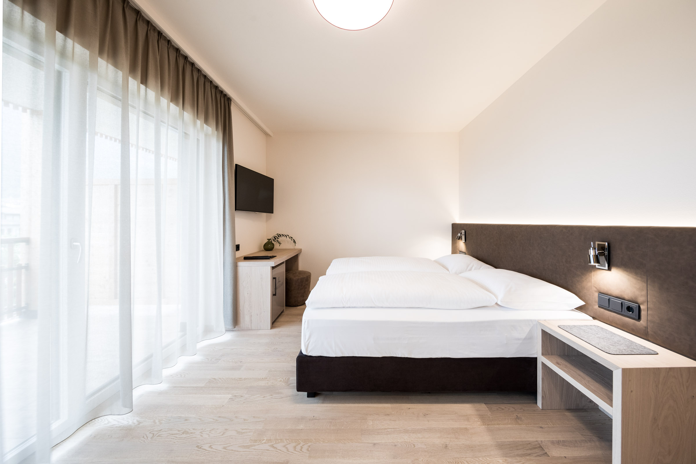 Modernes, schlicht eingerichtetes Schlafzimmer mit Doppelbett, hellem Holzboden und großen Fenstern mit Gardinen