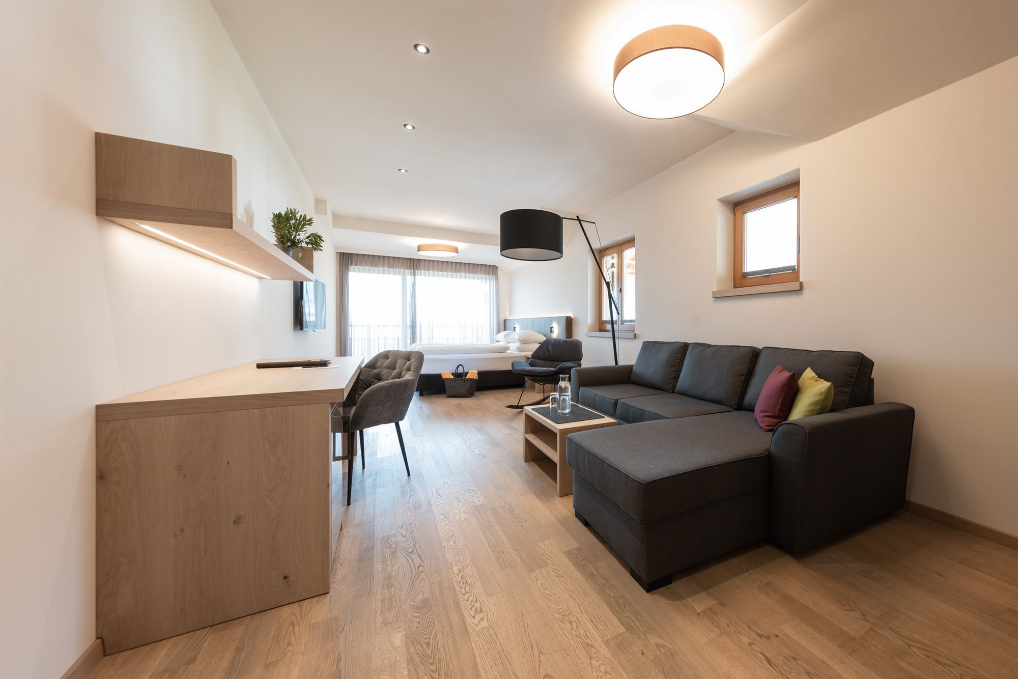 Modern eingerichtetes Wohnzimmer mit grauem Sofa, Schreibtisch und Bett im Hintergrund