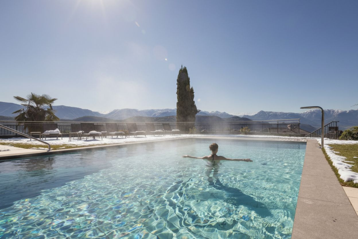 Person schwimmt im beheizten Außenpool mit Bergblick und Schnee am Rand
