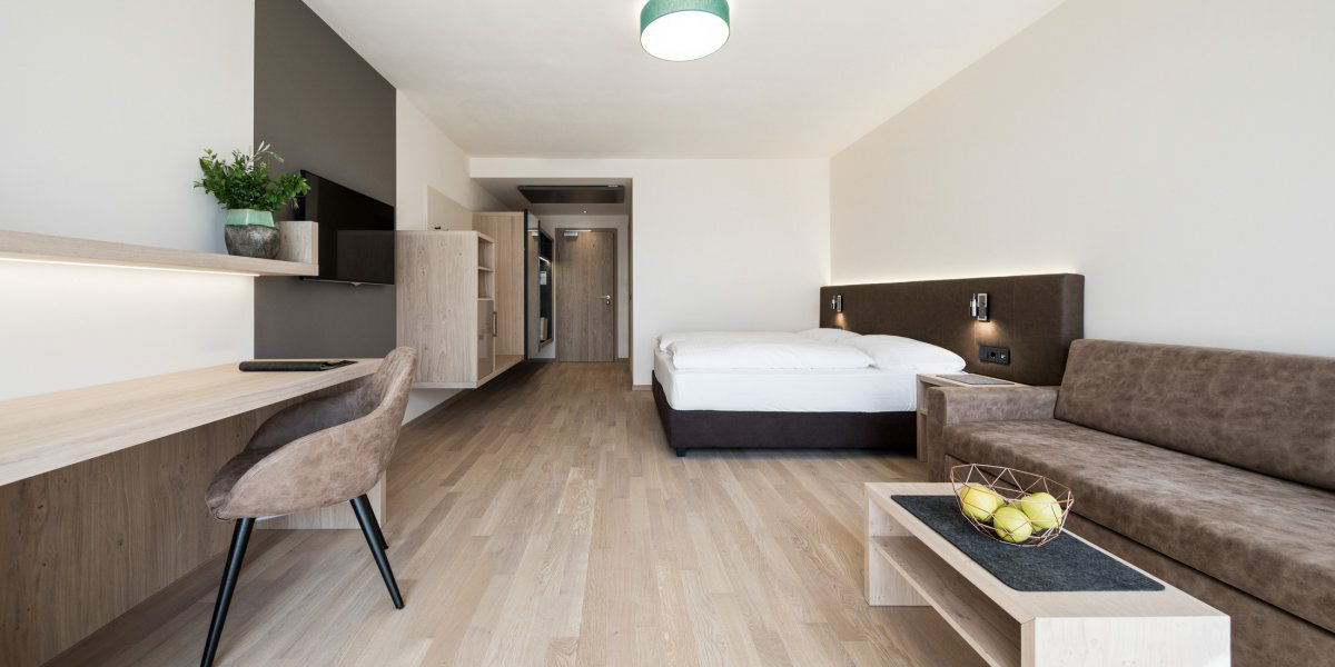 Modern hotel room with wooden floor, double bed, brown sofa, desk, and wall-mounted TV.