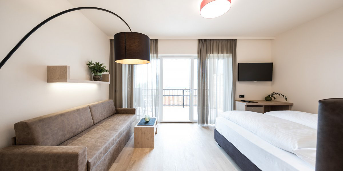 Modern hotel room with brown sofa, bed, small table, and balcony door with sheer curtains.